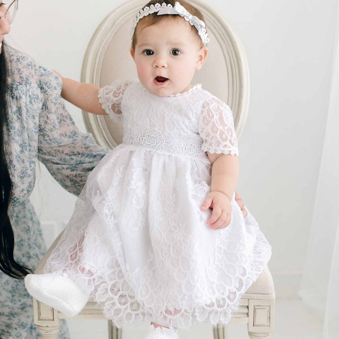 A toddler in an Olivia Dress is held by her mother while sitting in an antique chair, both facing away from the camera in a softly lit boutique room.