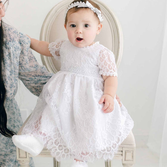 A toddler in an Olivia Dress is held by her mother while sitting in an antique chair, both facing away from the camera in a softly lit boutique room.