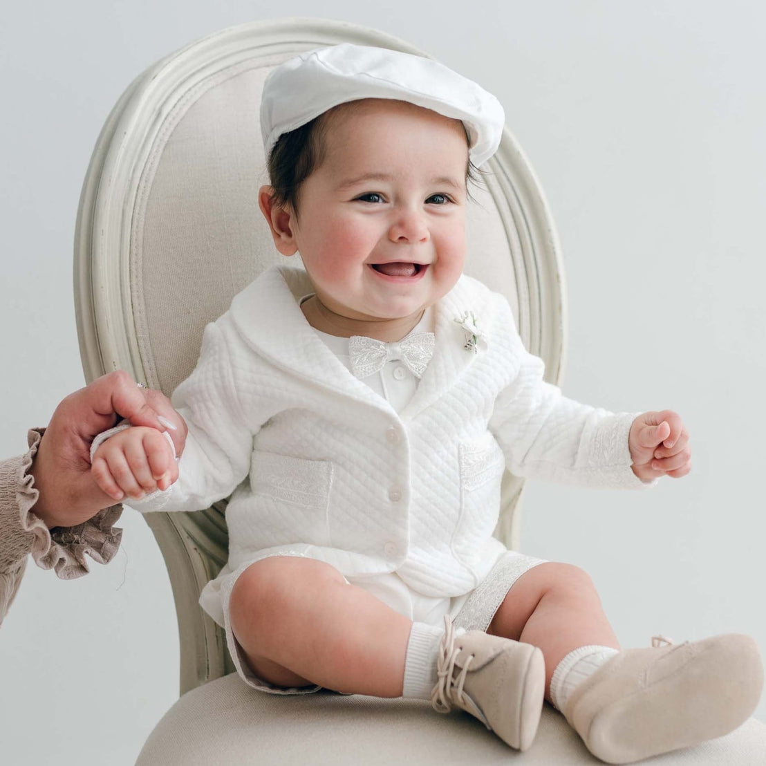 Smiling baby in the Oliver Quilted Jacket Suit with shorts, seated on a chair and holding an adult’s hand.