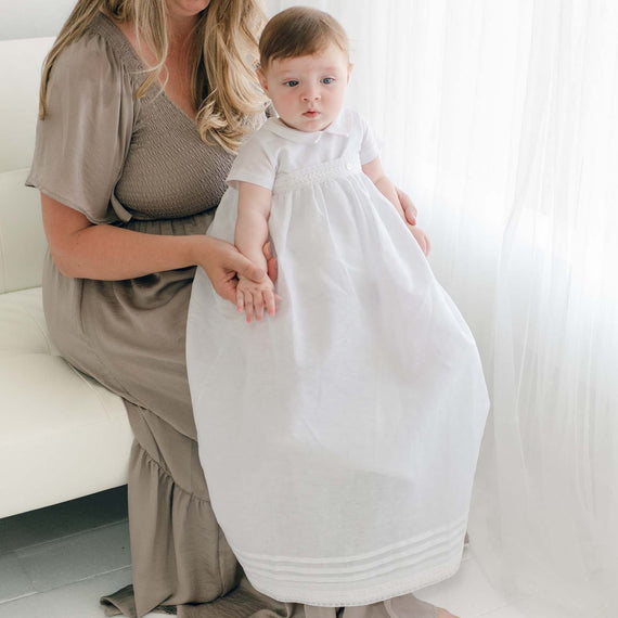 A woman with long hair, wearing a taupe dress, is sitting on a white couch. She is gently holding a baby dressed in an Oliver Convertible Gown | Romper & Skirt Set, who is standing on the floor. Natural light streams through a sheer curtain in the background.