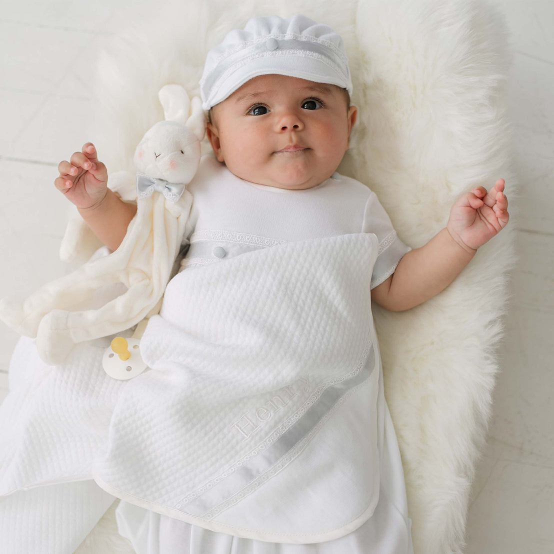 Baby in white outfit with a plush toy on a white blanket.