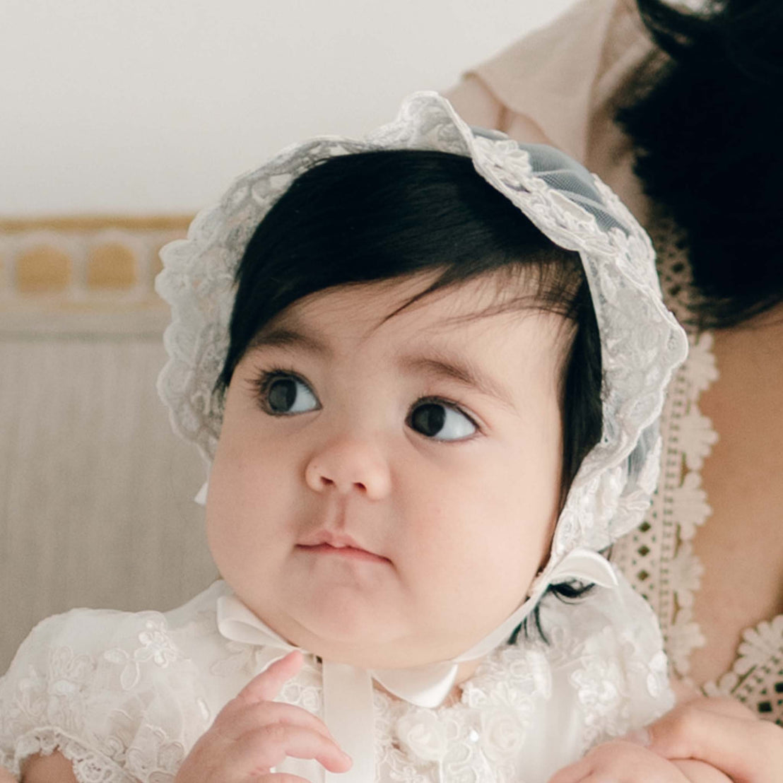 Baby girl wearing the Penelope Lace Bonnet and matching gown sits on the lap of her mother and looks away from the camera.
