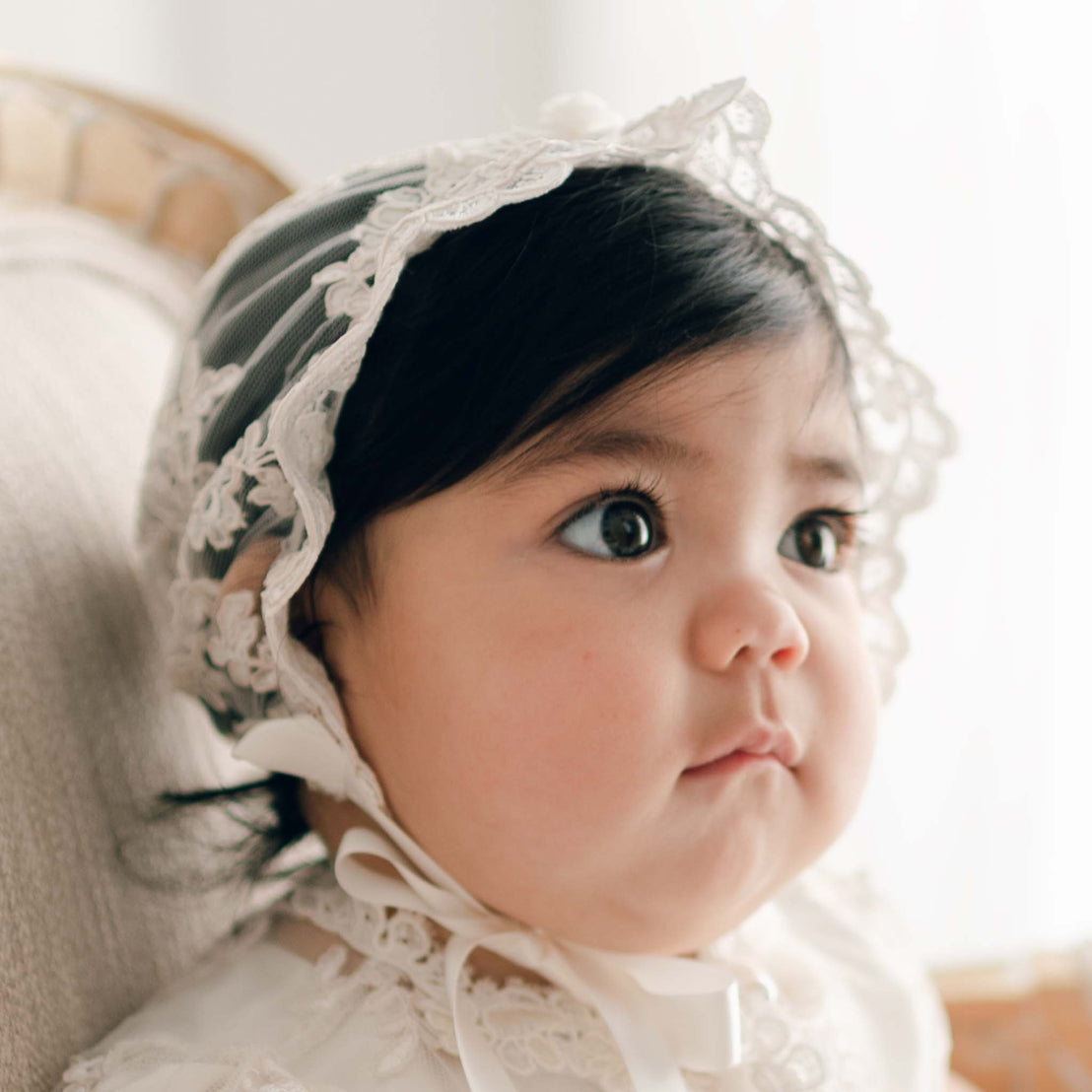 Baby girl with dark hair wearing the Penelope Lace Bonnet and matching baptism dress gazes away to the side of the camera.