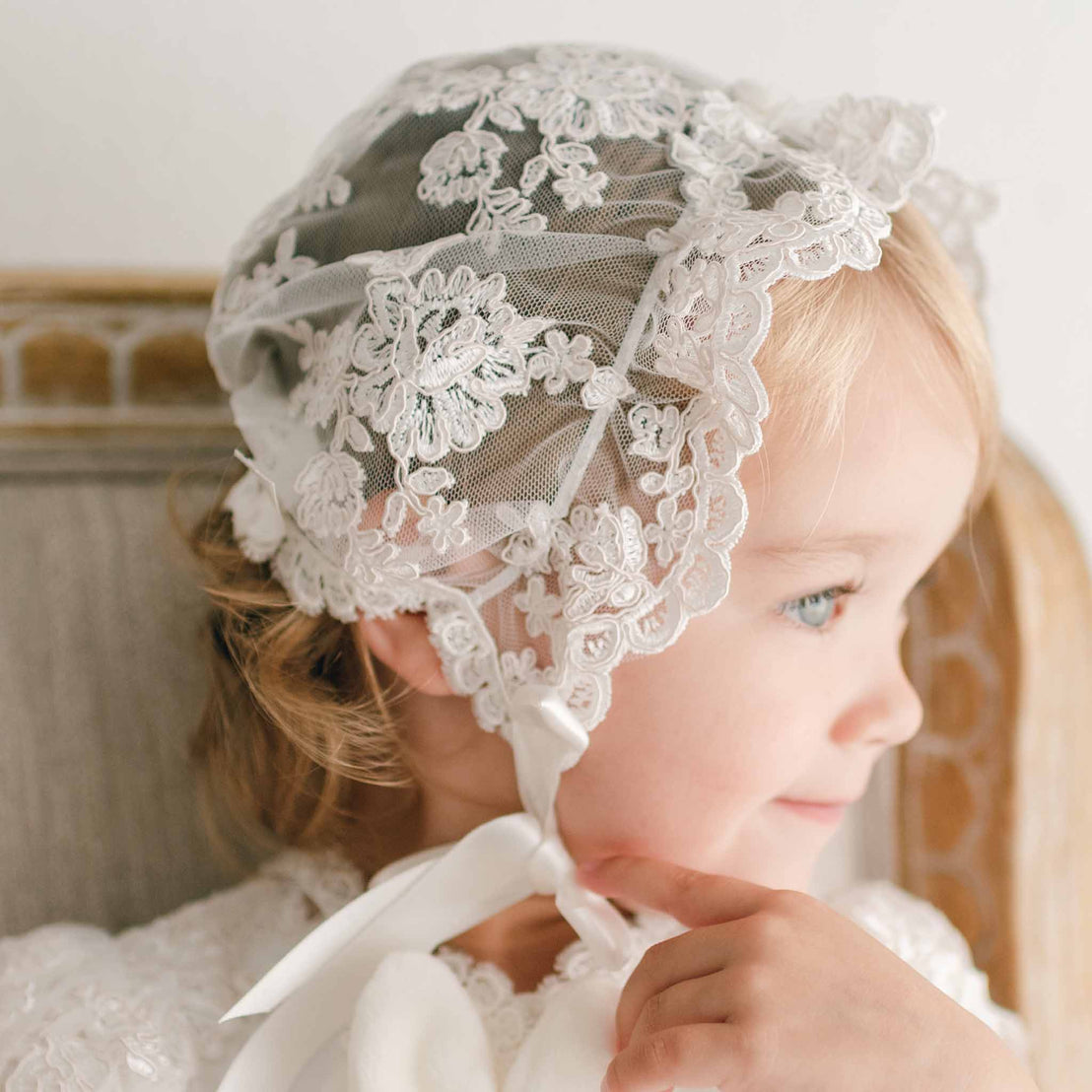 Close up detail of a baby girl slightly smiling in profile sitting in a beige upholstered chair and wearing the Penelope Lace Bonnet and matching gown.