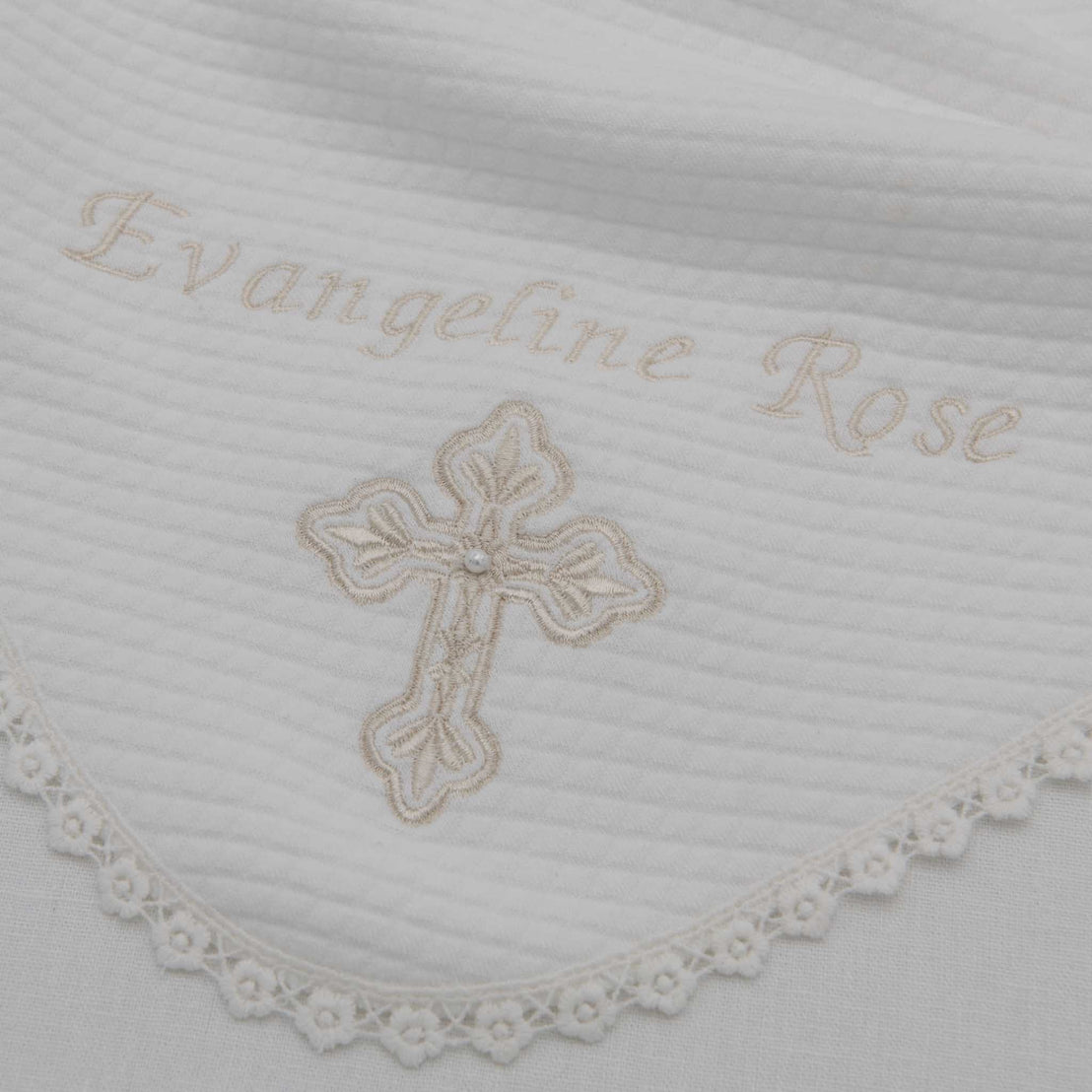 A close-up of a white fabric with the embroidered name "evangeline rose" and a delicate gold cross for a christening, surrounded by a lace trim edge on the Personalized Cross Blanket.