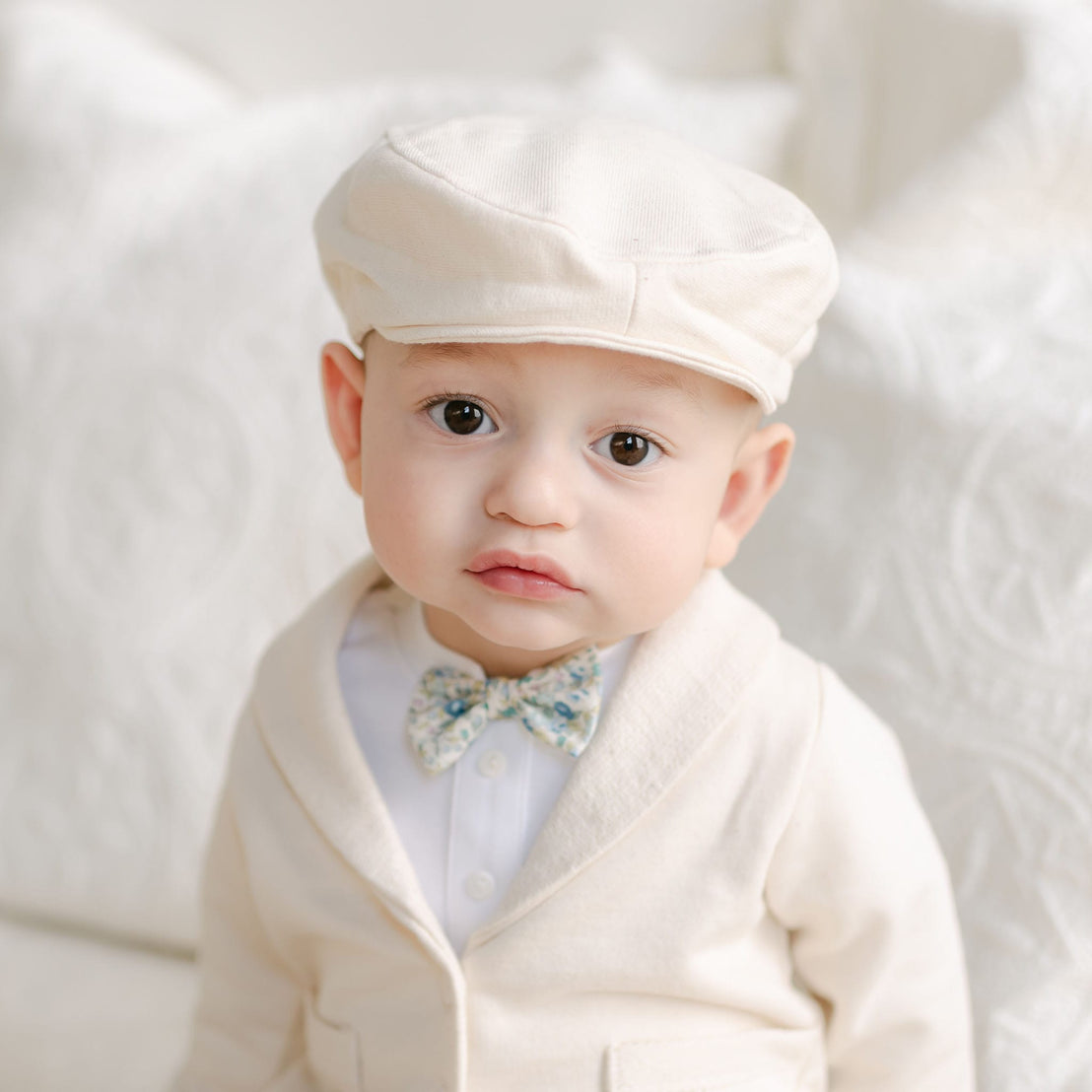 Baby wearing the Petite Fleur Blue Bow Tie and ivory cap with matching jacket.