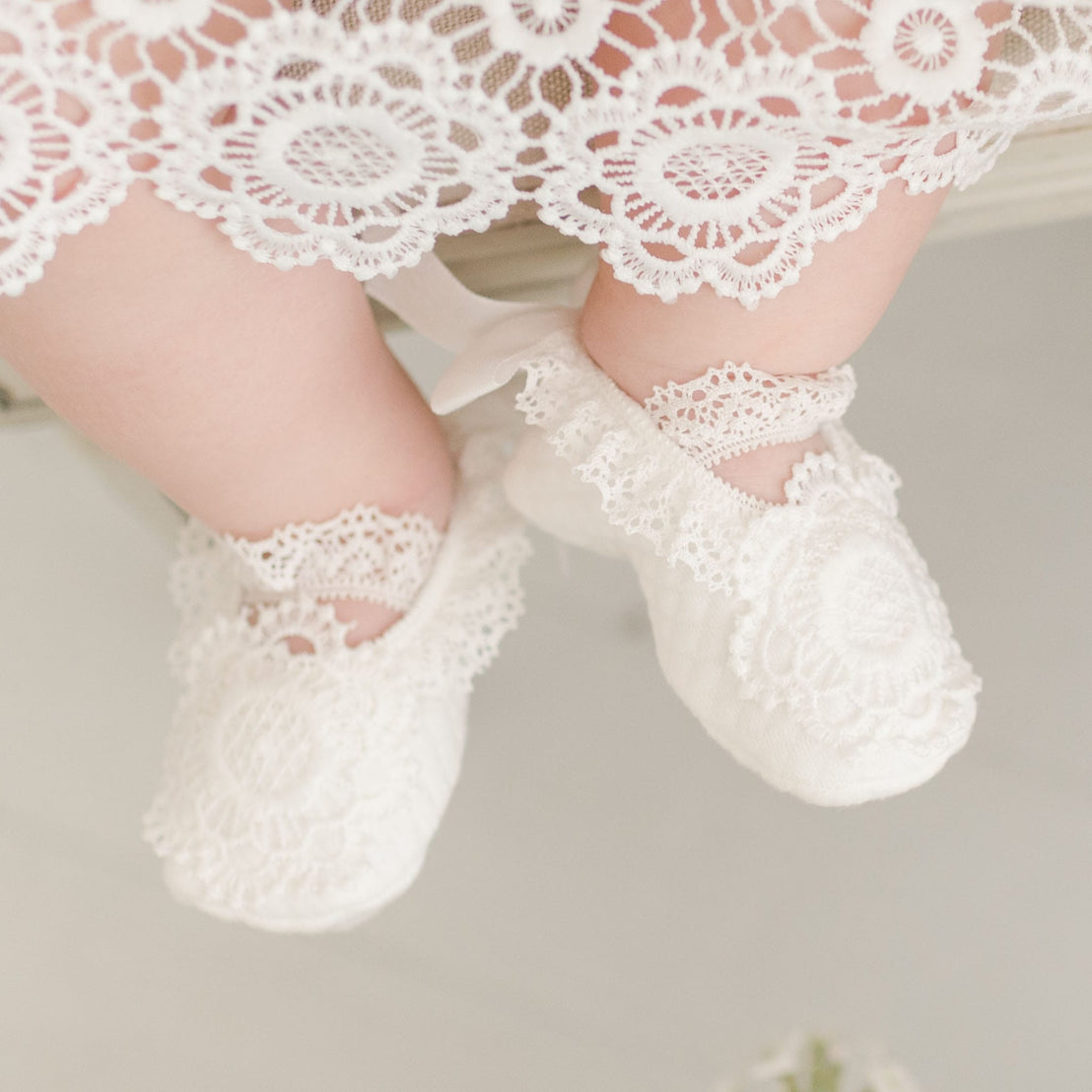 Baby feet in the Poppy Booties with intricate floral lace details, positioned under the lace hem of the Poppy Dress, resting gently on a neutral background.