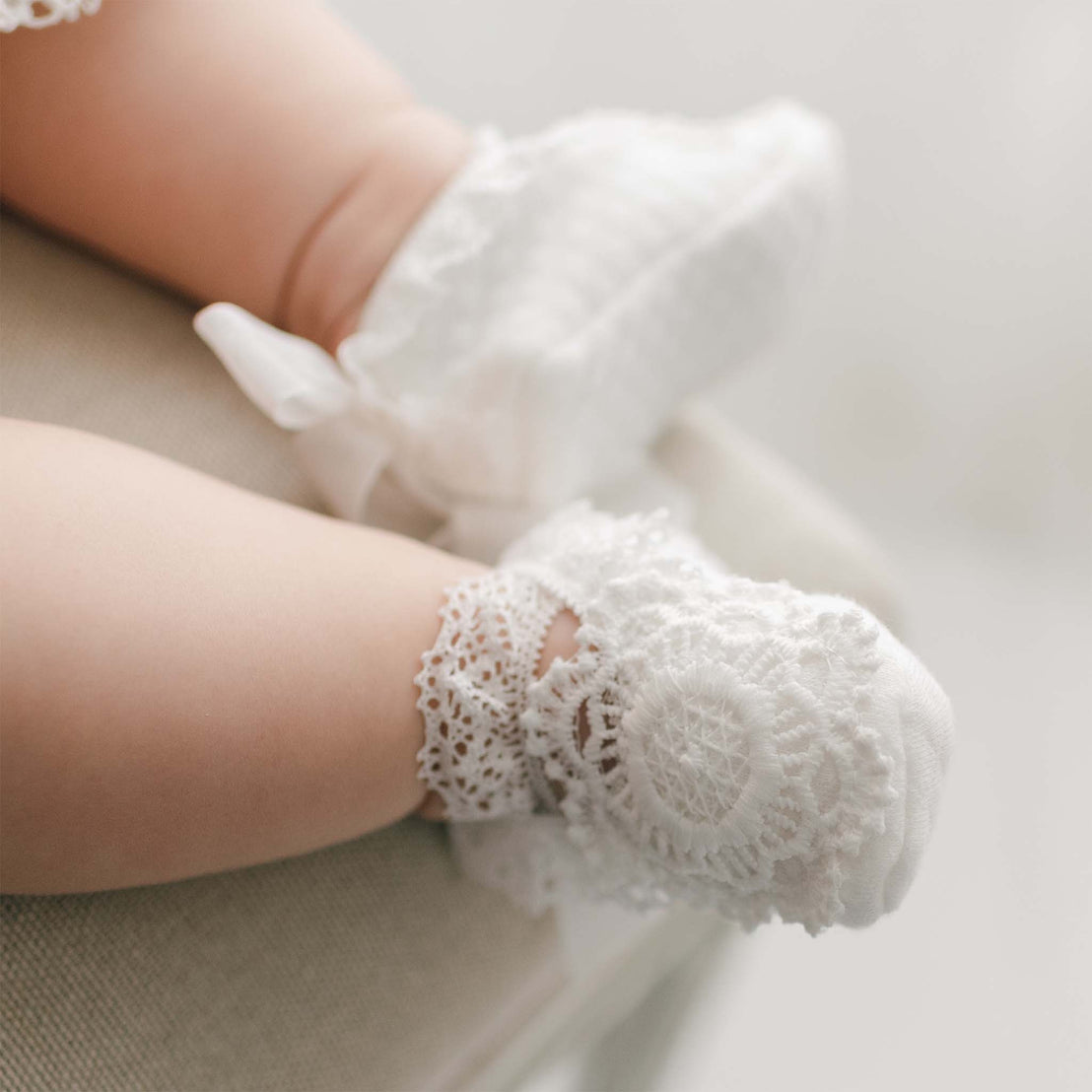 Close-up of baby feet wearing the Poppy Booties, featuring delicate floral lace and soft white fabric with ribbon ties, resting gently on a cream chair.