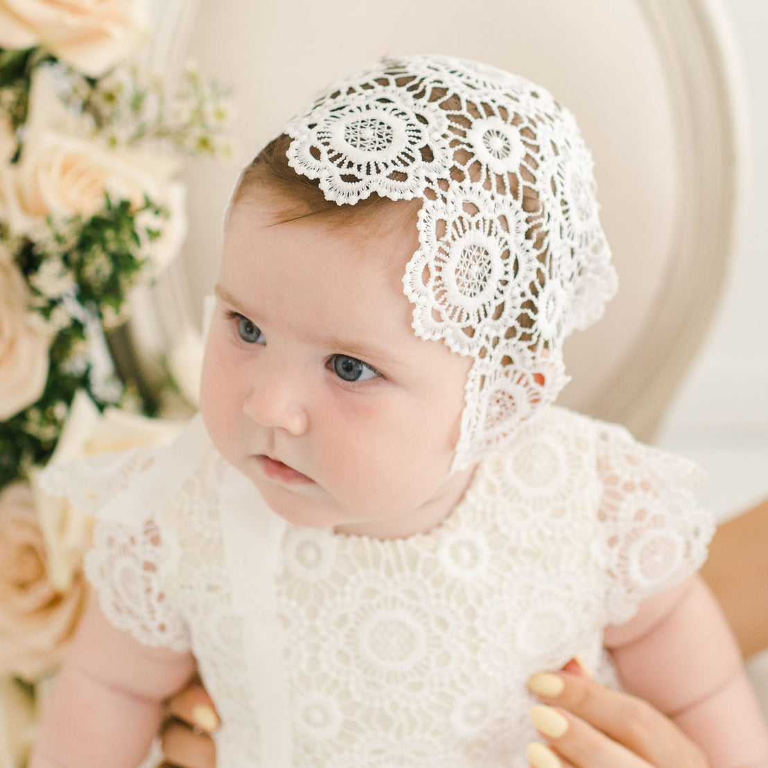 Baby wearing the Poppy Christening Gown and matching lace bonnet, showcasing the intricate floral lace design and soft ivory fabric, seated in front of a neutral chair with light peach roses in the background.