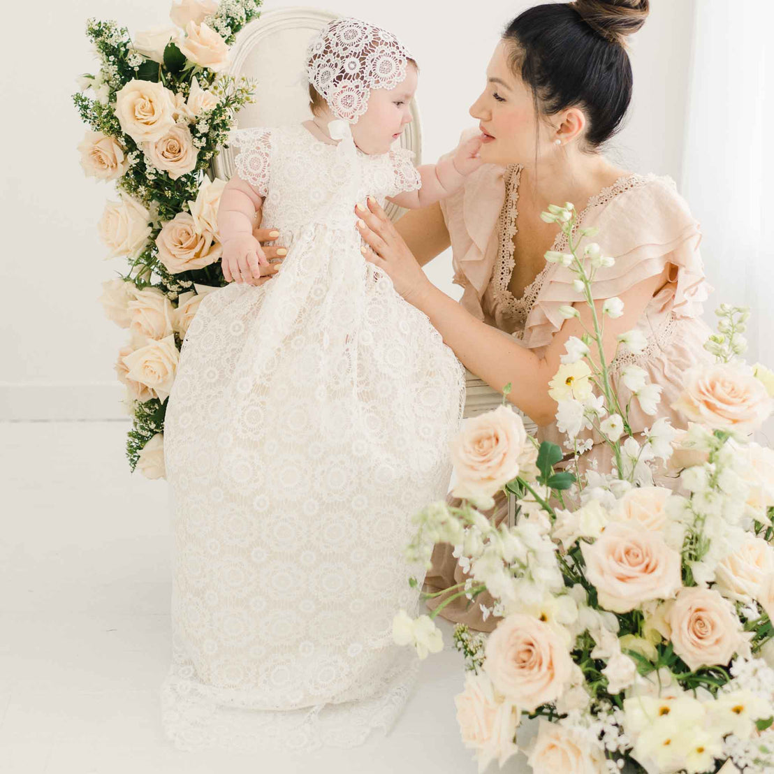 Baby girl in the Poppy Christening Gown and Bonnet, sitting on a white chair surrounded by floral arrangements, interacting with her mother, who is kneeling beside her.