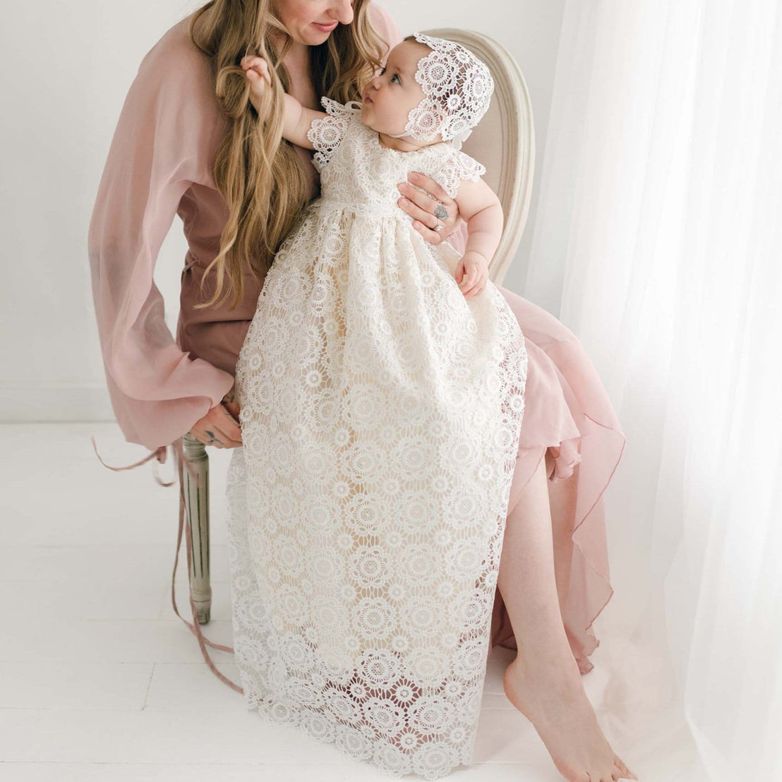 Baby wearing the Poppy Christening Gown and Bonnet, sitting on a woman's lap, showcasing the intricate floral lace overlay and long gown design in soft natural light.