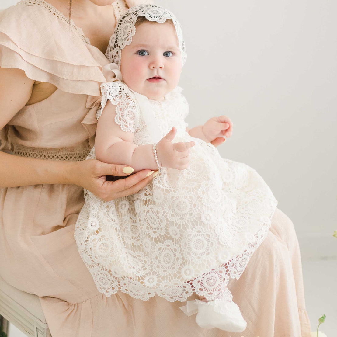 A baby girl wearing the Poppy Lace Dress and matching bonnet is held by her mother. The intricate lace design and soft ribbon tie on the bonnet are highlighted.