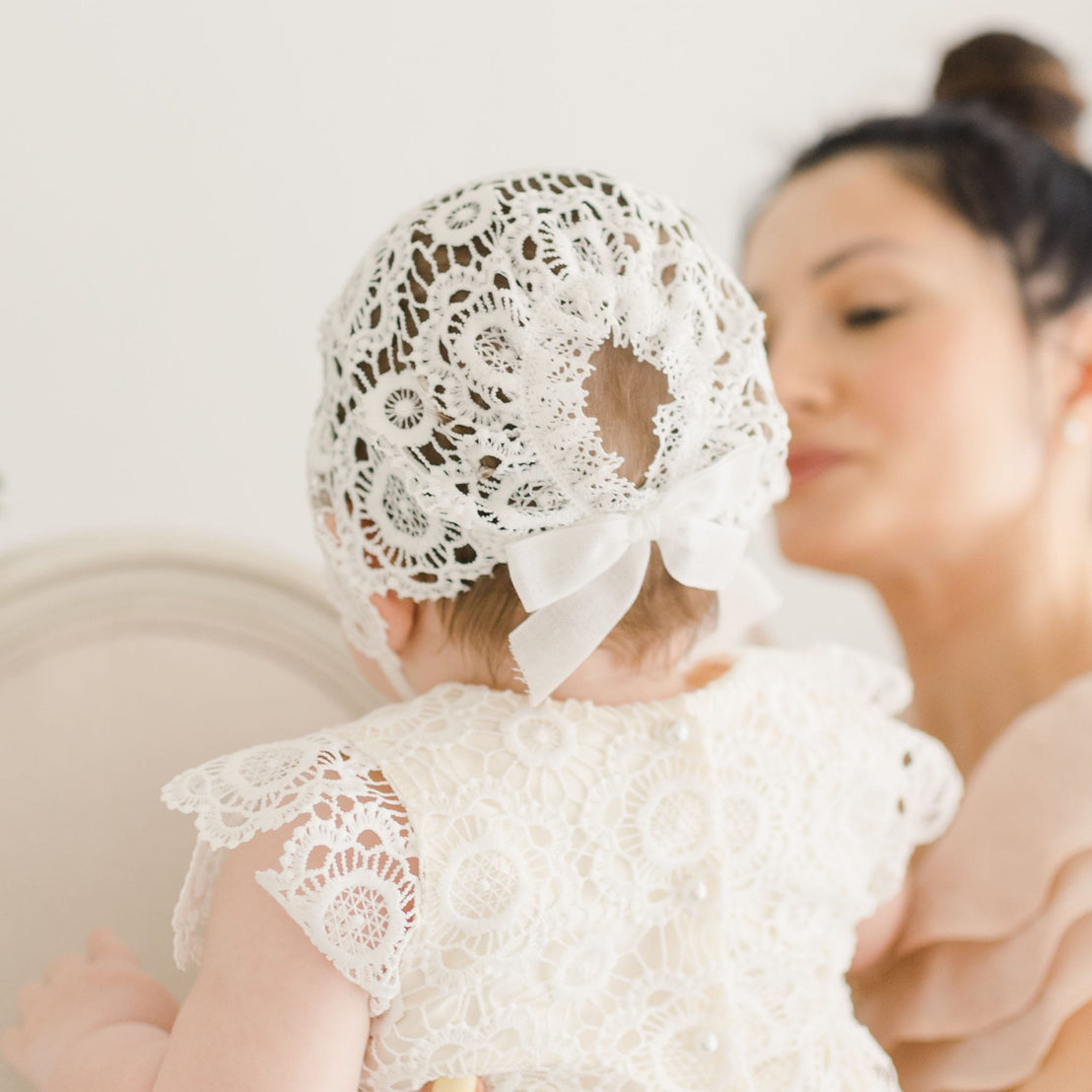 Back view of the Poppy Dress and Bonnet, highlighting the bonnet's intricate floral lace and satin bow detail as the baby girl is held by her mother.