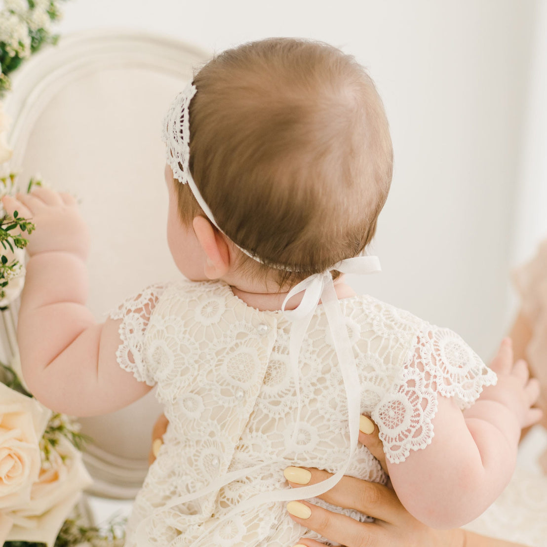 Back view of a baby wearing the Poppy Lace Headband with a white ribbon tied at the back, paired with the matching dress adorned with pearl button details.