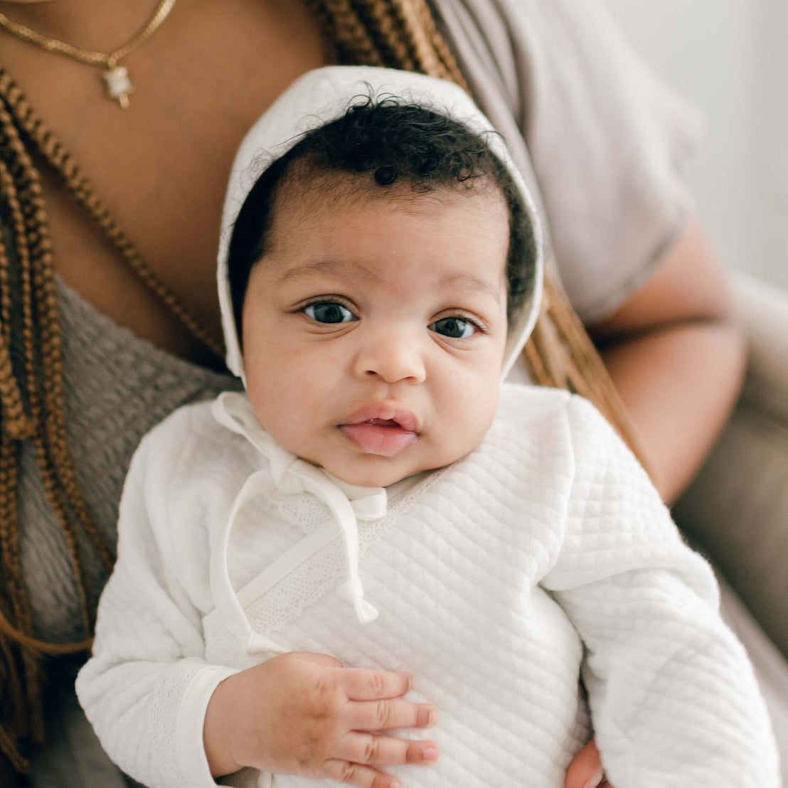 Baby girl with her mother and wearing the Ivory Quinn Quilted Cotton Bonnet. A traditional style bonnet that is lined in coordinating super soft pima cotton and trimmed in a light ivory cotton lace with soft pima cotton ties to secure the bonnet.