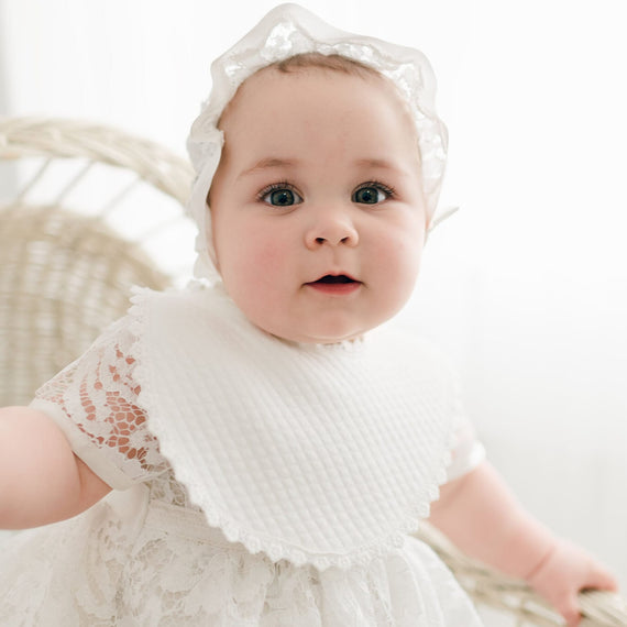 A baby with blue eyes and a Rose Bib of ivory Venice floral lace, with a matching headband, sits in a wicker chair, looking curious and adorable.
