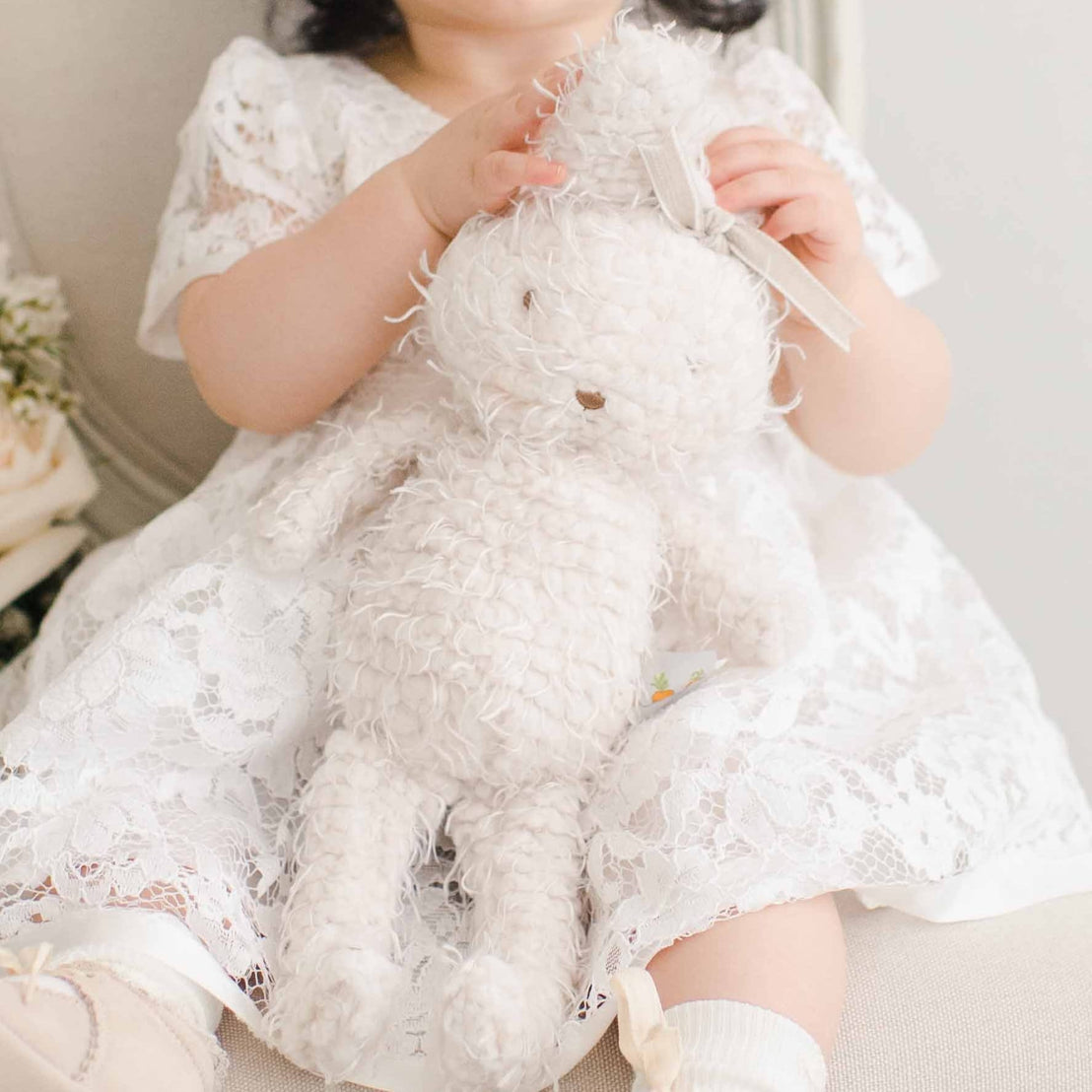 A toddler in a traditional white lace dress sits on a chair holding a white plush Rose Bunny, with a vintage bouquet of soft pink roses to the side.