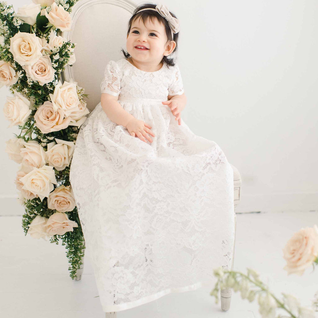 A cheerful toddler girl in a Rose Christening Gown & Bonnet sits on an elegant chair decorated with pale pink roses, in a bright, upscale room.