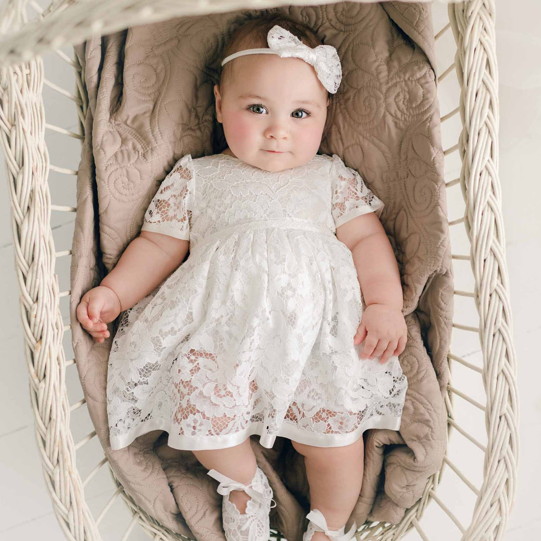 A baby dressed in the Rose Romper Dress and a matching Rose Bow Headband, lies comfortably in a wicker basket, looking up towards the viewer with a gentle expression.