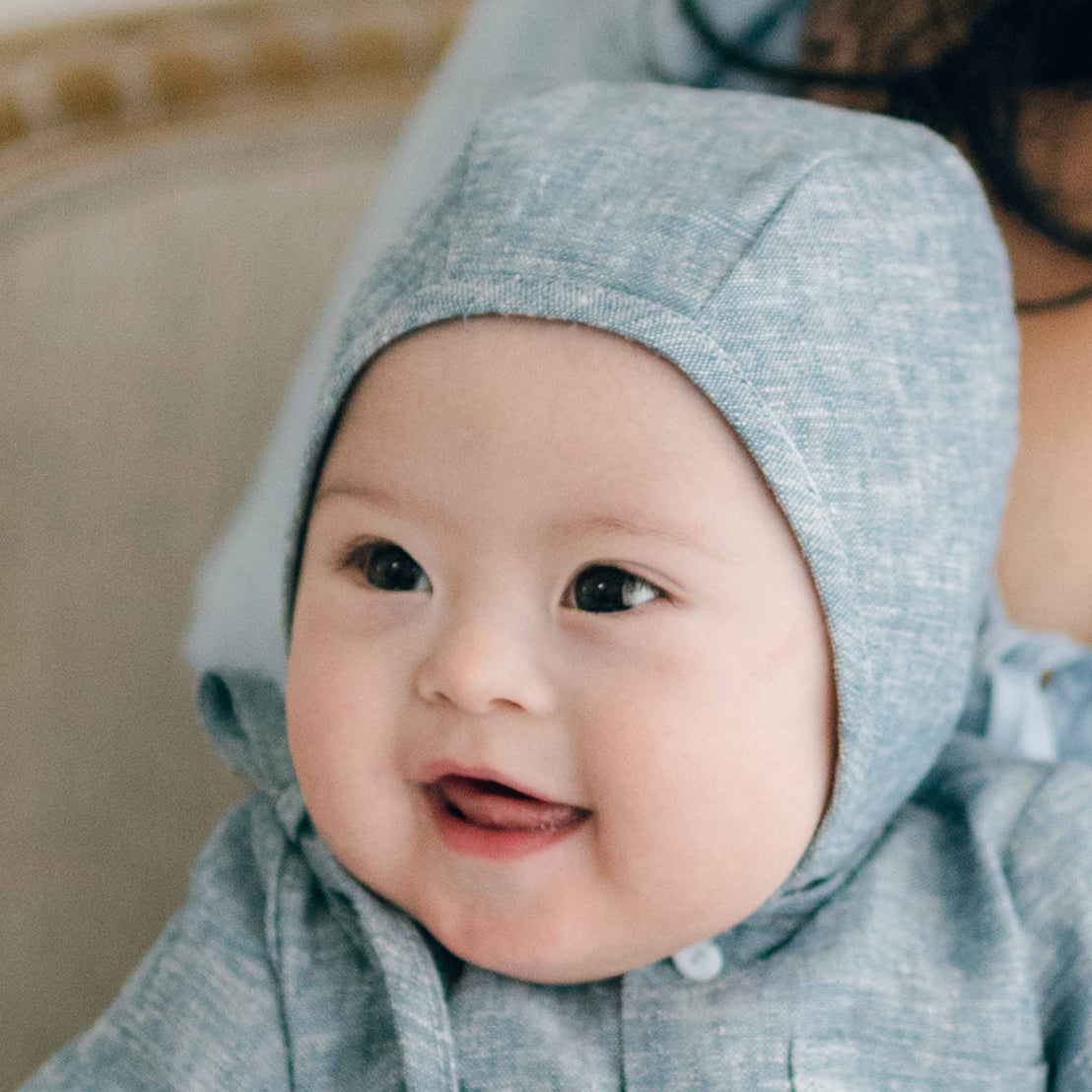 Wearing a Henry Linen Bonnet, the smiling baby with rosy cheeks and wide eyes gazes off to the side, sitting comfortably in a softly lit indoor setting.