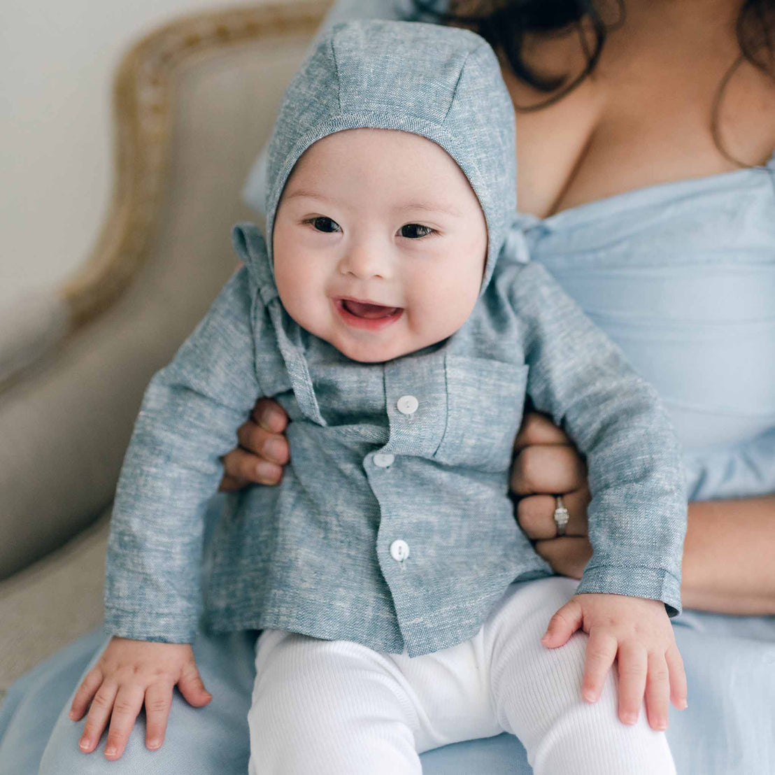 A smiling baby wearing a Henry Linen Shirt and matching hat sits on a woman's lap. The woman, dressed in a light blue dress, gently cradles the baby as they both rest on a light-colored chair, enveloped in the cozy and joyful ambiance of handmade charm.