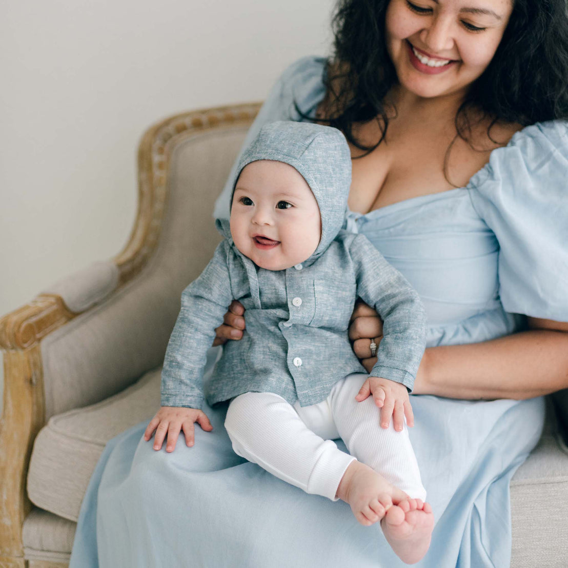 A smiling woman in a light blue Henry Linen Shirt cradles a baby who is dressed in a handmade hooded outfit and white pants. They are seated on a beige chair, with the baby appearing happy and playful.