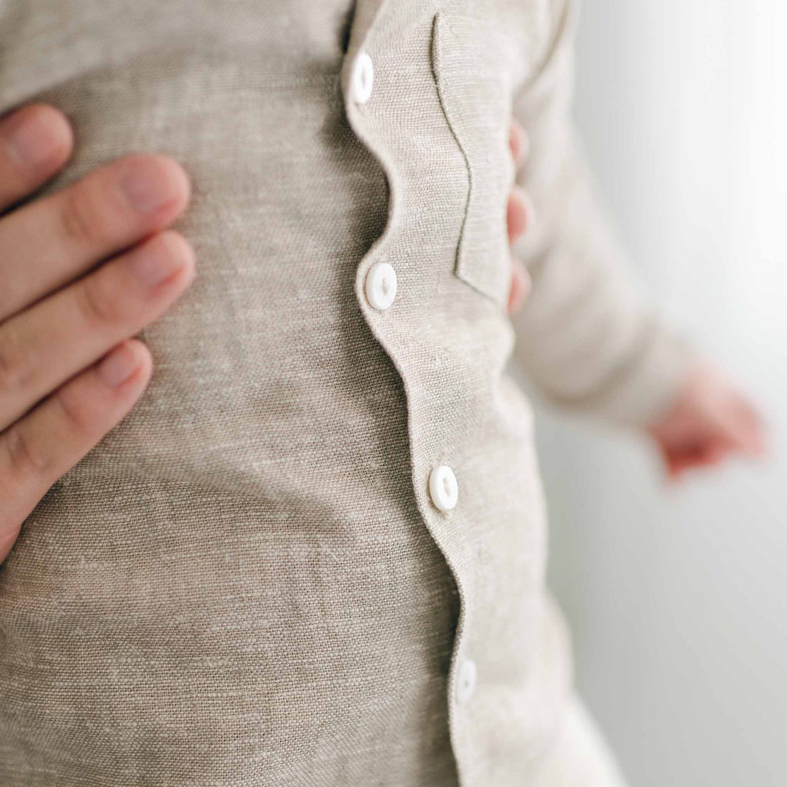 A close-up of a person gently cradling a baby dressed in the Henry Linen Shirt, a light beige outfit made from a linen-cotton blend and adorned with white buttons. The image beautifully conveys the tactile texture of the fabric along with the warm, nurturing gesture.