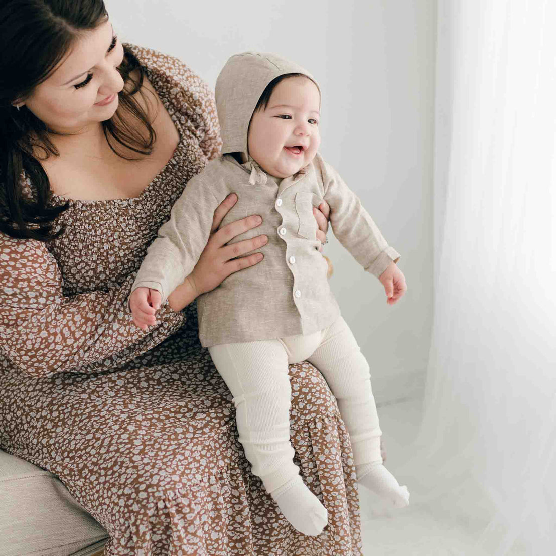 A woman in a floral dress cradles a smiling baby wearing the cozy, handmade Henry Linen Shirt. The softly lit scene with its light background creates a warm and inviting atmosphere.