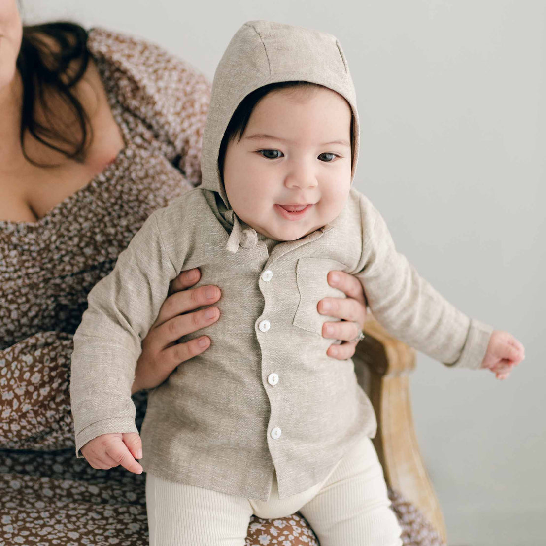 A baby, dressed in a Henry linen shirt and white pants made from a linen/cotton blend, is being held by a woman wearing a floral dress. The happy child sits on a wooden chair set against a light background.