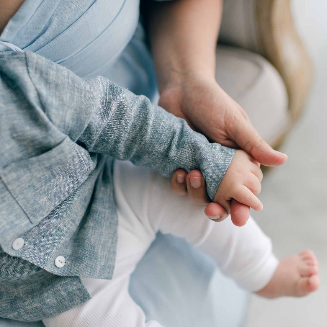 A close-up of an adult holding a baby's hand. The baby wears a gray long sleeve shirt paired with the Henry Linen Shirt Set in French terry cotton pants, while the adult is dressed in a blue outfit. The indoor setting, with its soft lighting, creates a calm atmosphere perfect for this tender moment.