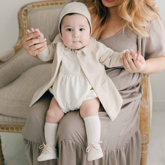 A baby in a vintage-style beige outfit made from French terry cotton, complete with a bonnet and knee-high socks, is perched on a woman's lap. The woman is wearing the Henry Trench Coat in taupe as she gently holds the baby's hands while they sit on a plush chair.