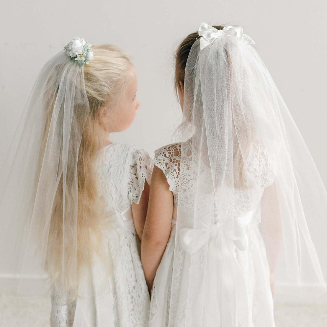 Two girls wearing the Veil, each attached with a floral crown or silk bow, draping softly over their lace dresses.
