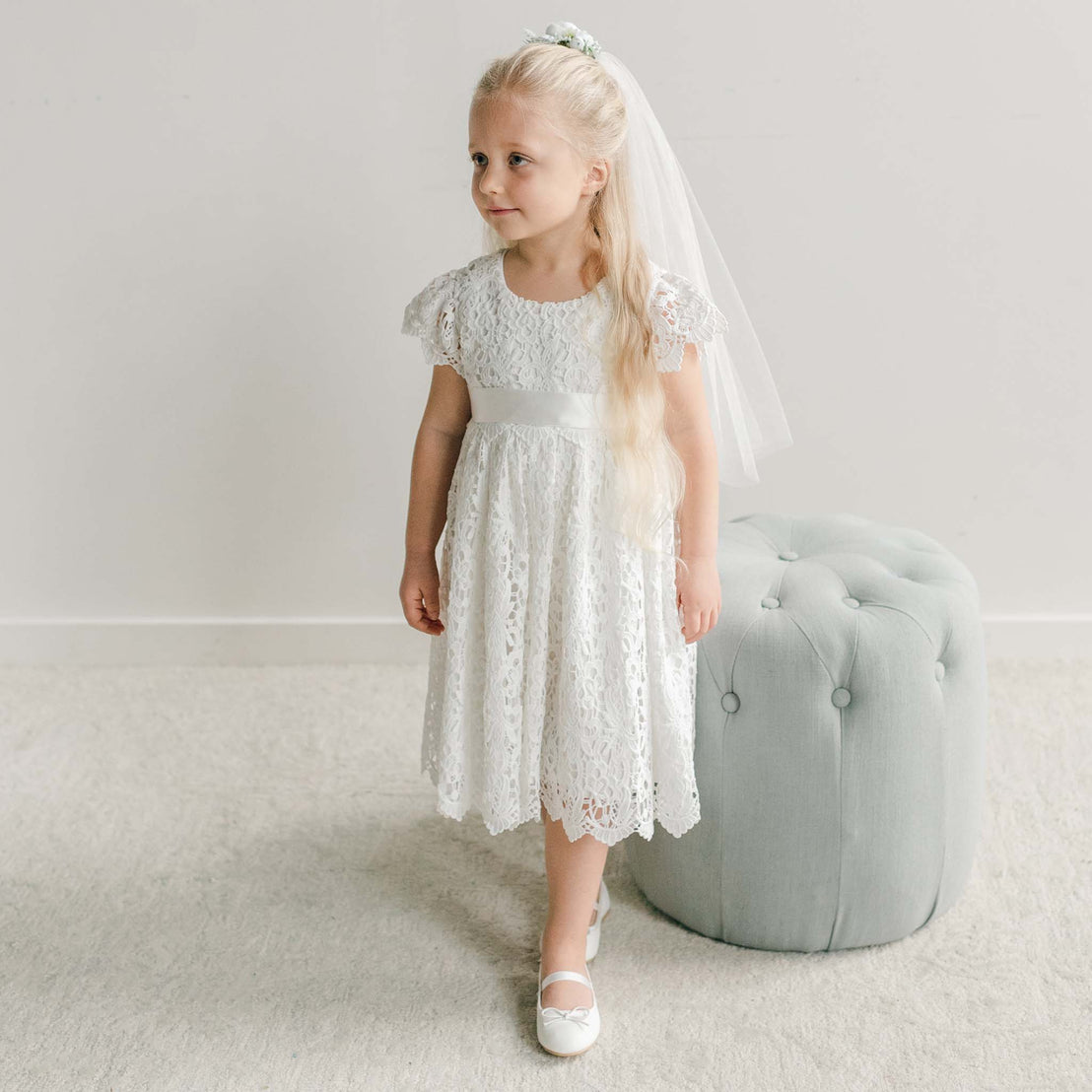 A girl wearing a white silk ribbon sash with a tulle veil, styled with her long hair in a half-up look.