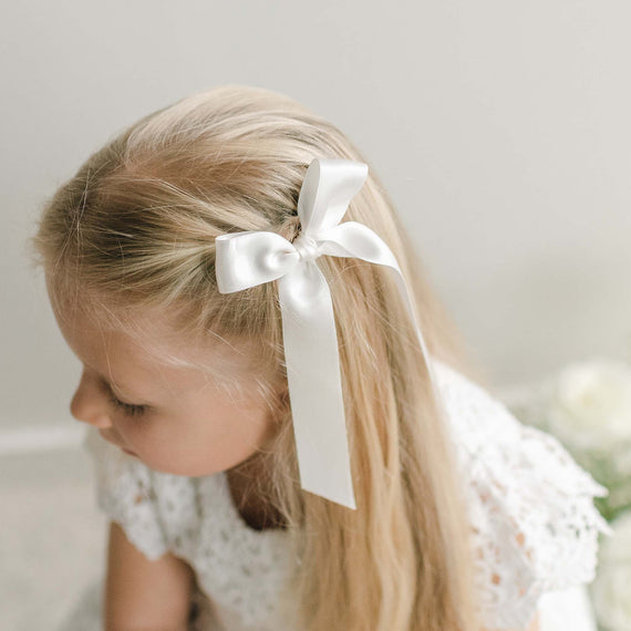 A white silk ribbon hair bow tied into a side style on a girl's long blonde hair.