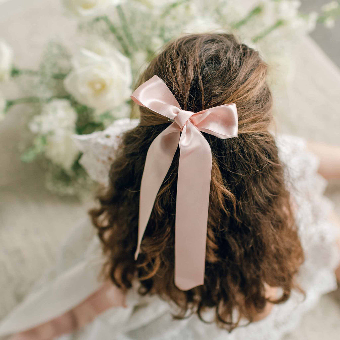 A close-up of a girl’s hairstyle adorned with a blush pink silk ribbon hair bow, styled alongside loose curls.