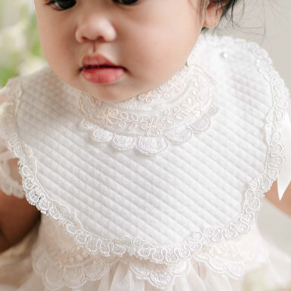 A baby wearing the Sophia Bib, sitting among soft white and blush flowers, dressed in a matching lace outfit and a coordinating headband.