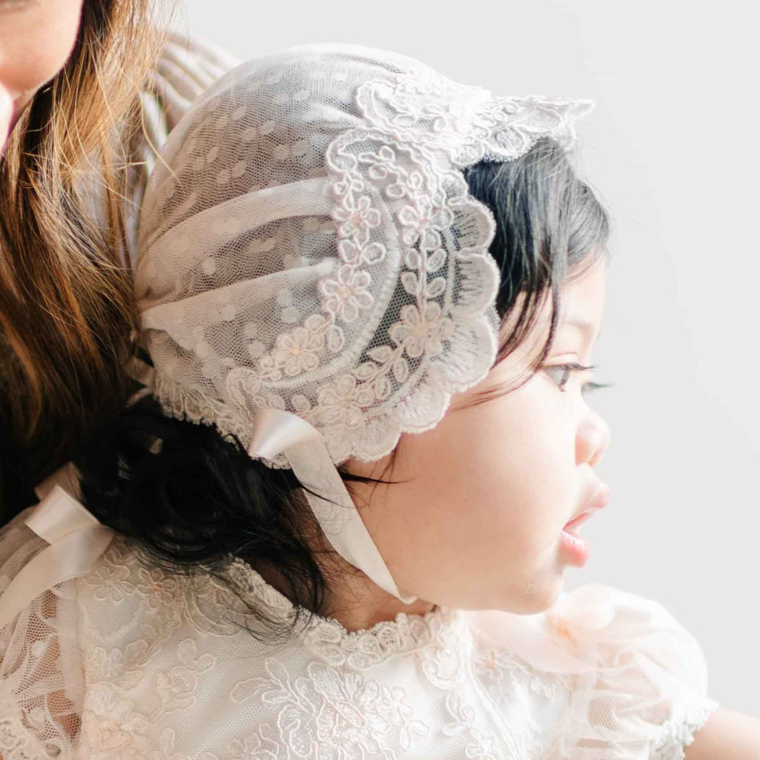 A close-up of a baby wearing the Sophia Bonnet, featuring delicate lace with floral embroidery and soft satin ribbon ties, paired with the Sophia Dress.