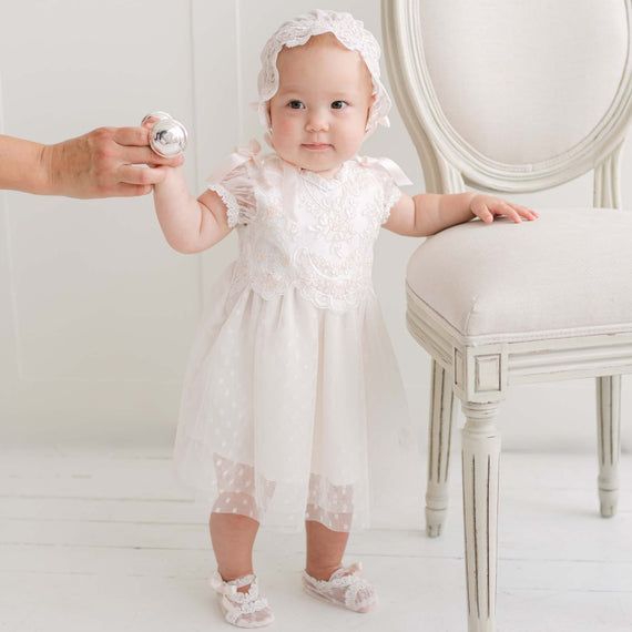 A baby wearing an ivory and pink lace and tulle baptism dress with a matching headband stands next to a white chair, holding a silver rattle held by an adult hand.