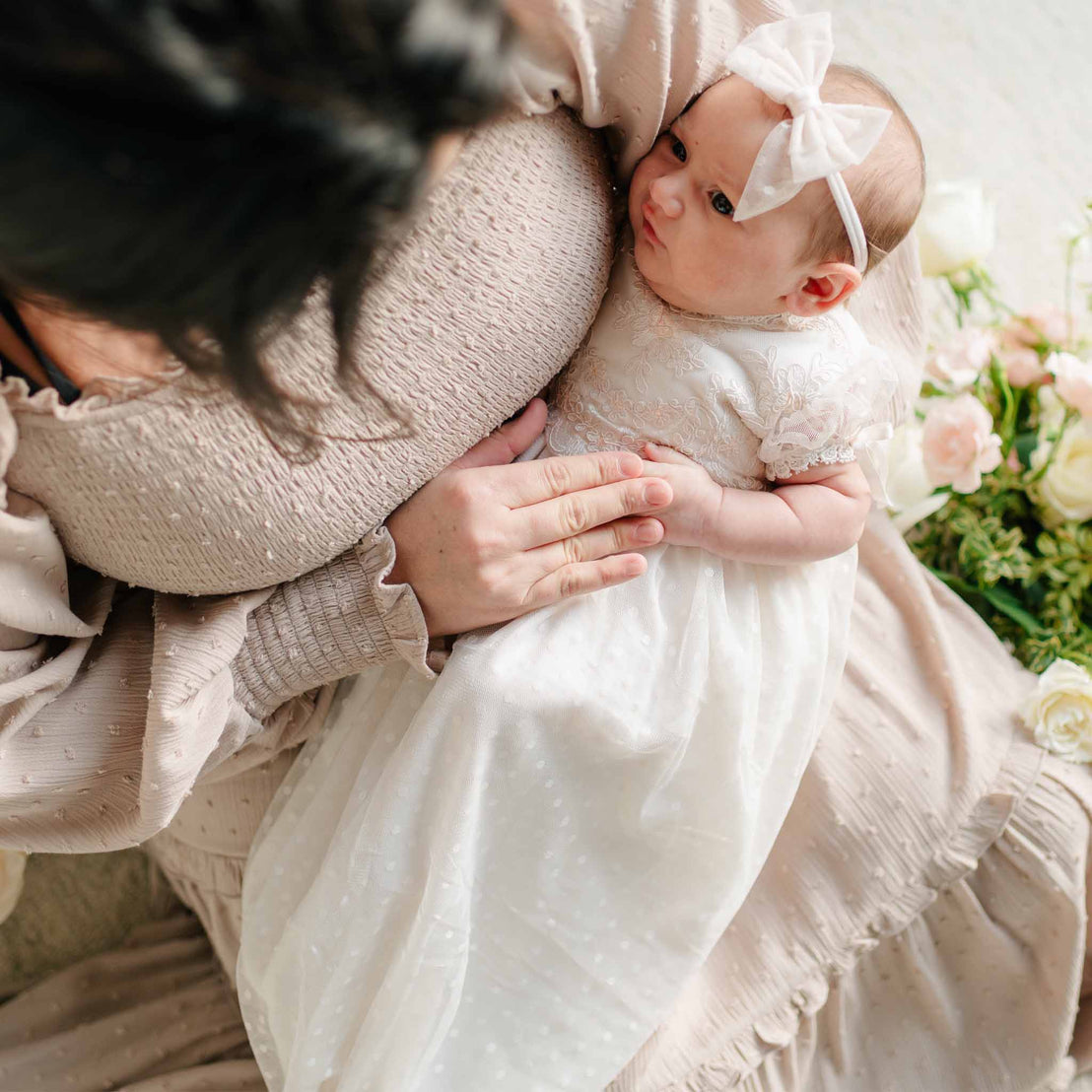 A newborn rests in their mother’s arms wearing the Sophia Layette, a delicate white gown with embroidered lace bodice and flutter sleeves, paired with a soft bow headband.