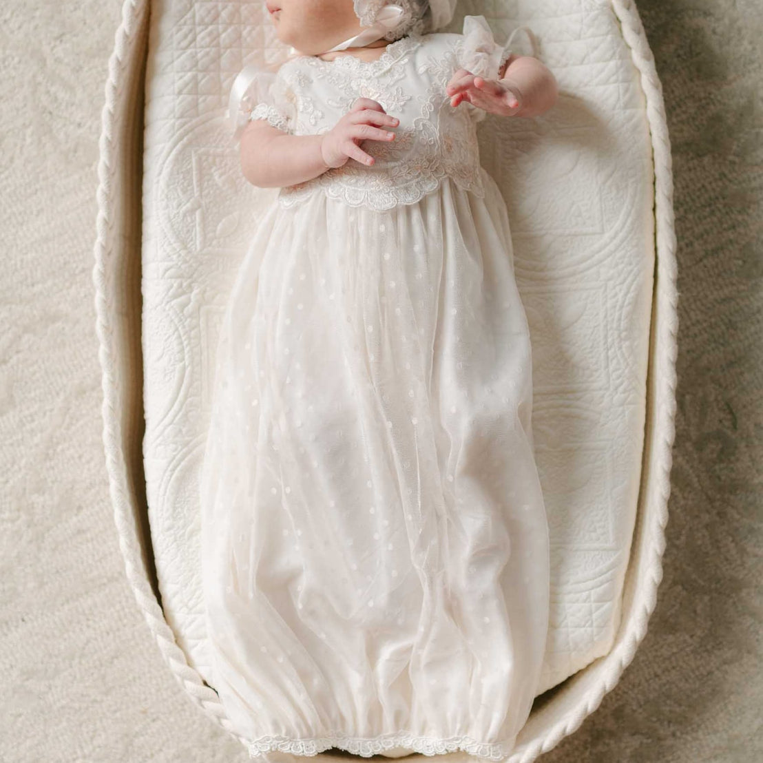 A newborn lays in a woven bassinet, dressed in the Sophia Layette with a matching lace bonnet, surrounded by soft textures.