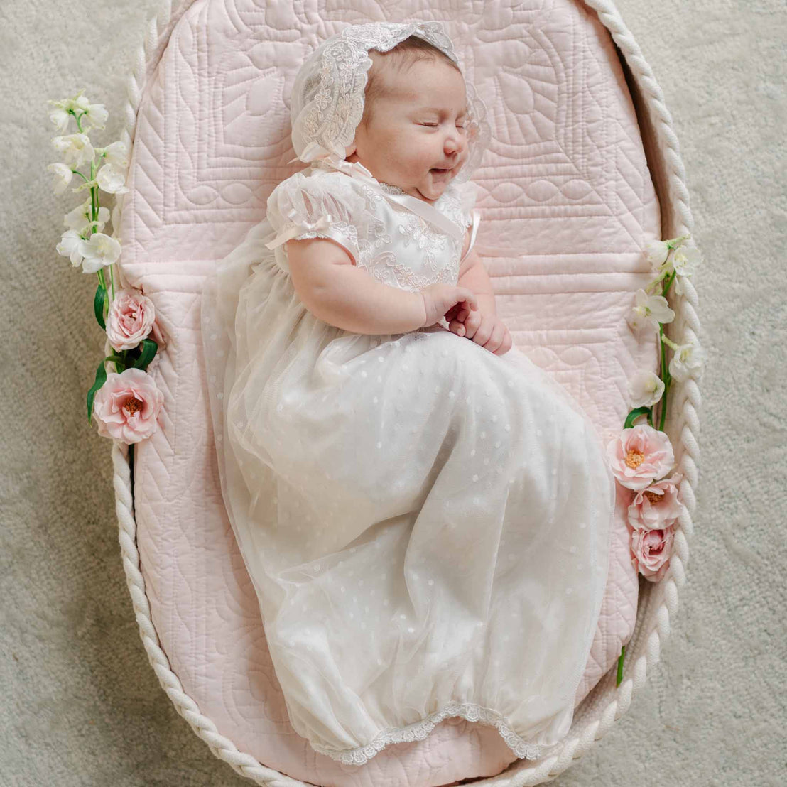 A baby sleeps in a bassinet adorned with flowers, dressed in the Sophia Layette with a lace bonnet and flutter sleeves.