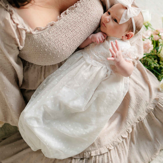 A close-up of a baby cradled in their mother’s embrace, highlighting the embroidered lace details and flowing dotted skirt of the Sophia Layette.