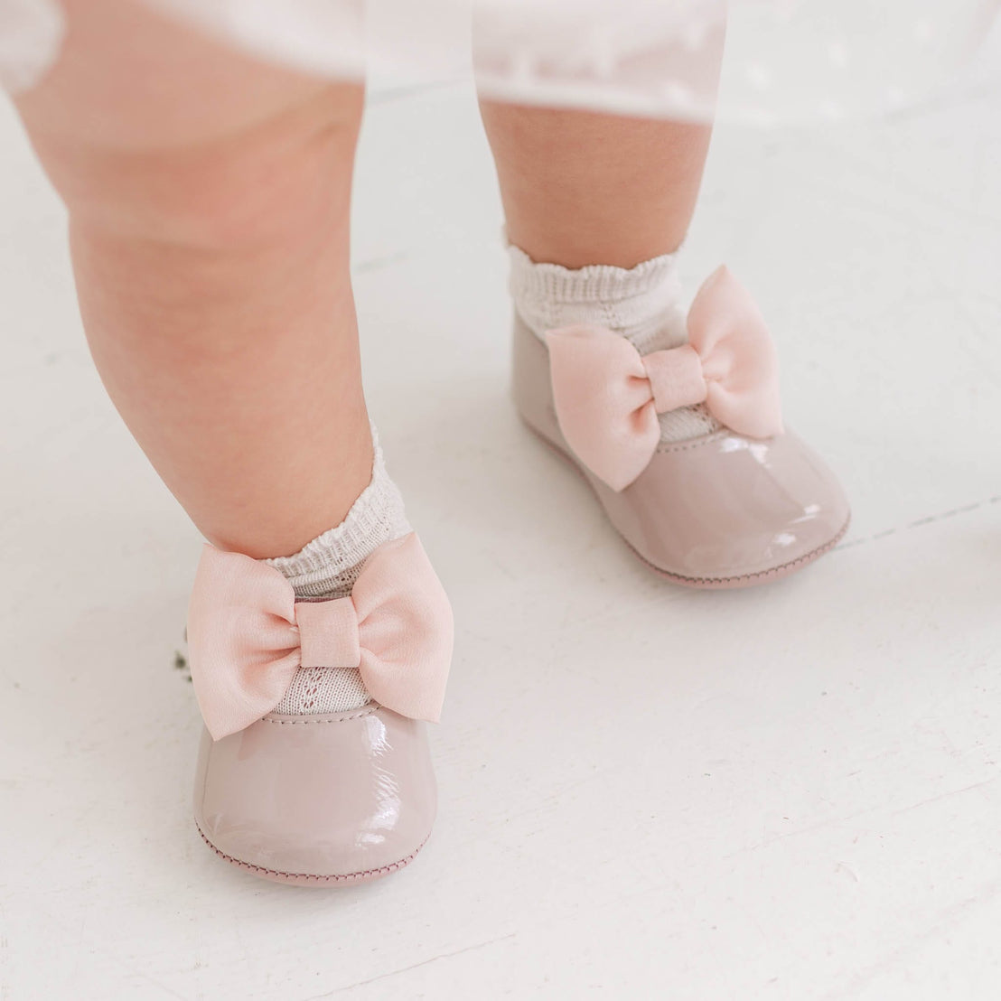 A close-up of a baby's feet wearing shiny pale pink patent leather mule shoes with large matching bow accents, paired with white ribbed socks, standing on a white floor.