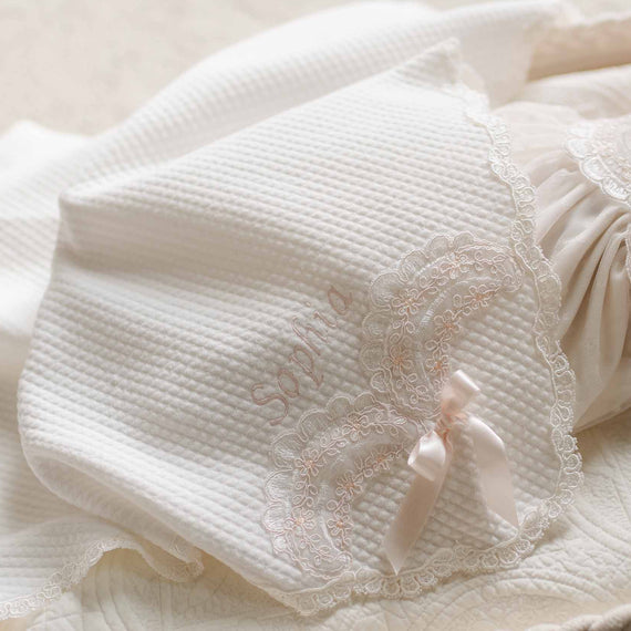 Close-up of the Sophia Blanket, featuring quilted white cotton with embroidered lace detailing, a delicate satin bow, and "Sophia" embroidered in soft blush thread.