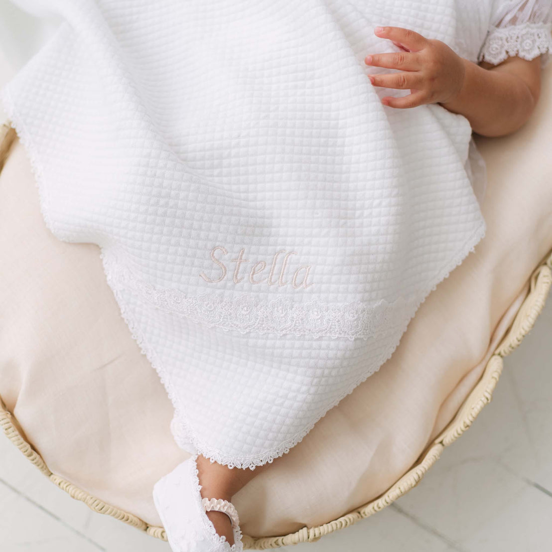 Close up photo of the Stella Personalized Blanket covering a baby in a wicker bassinet. Details show the corner of the blanket embroidered with the name "Stella" and trimmed with white edge lace. Part of Baby Beau & Belle's high end baby blankets.