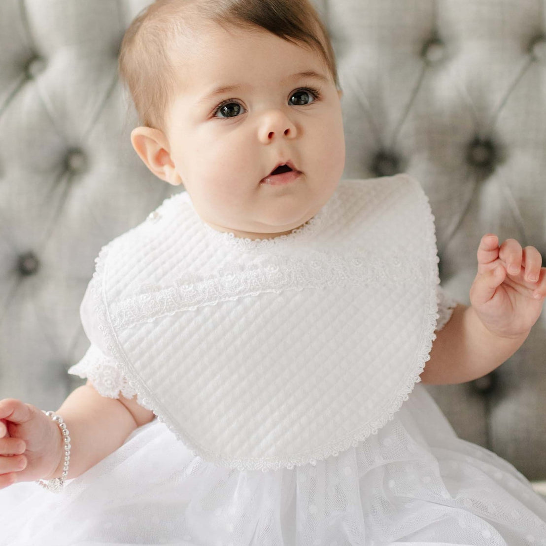 Baby wearing the Stella Bib against a gray tufted background. Details of the bib show a quilted cotton with a white edge lace trim. Perfect for unique baby gifts.