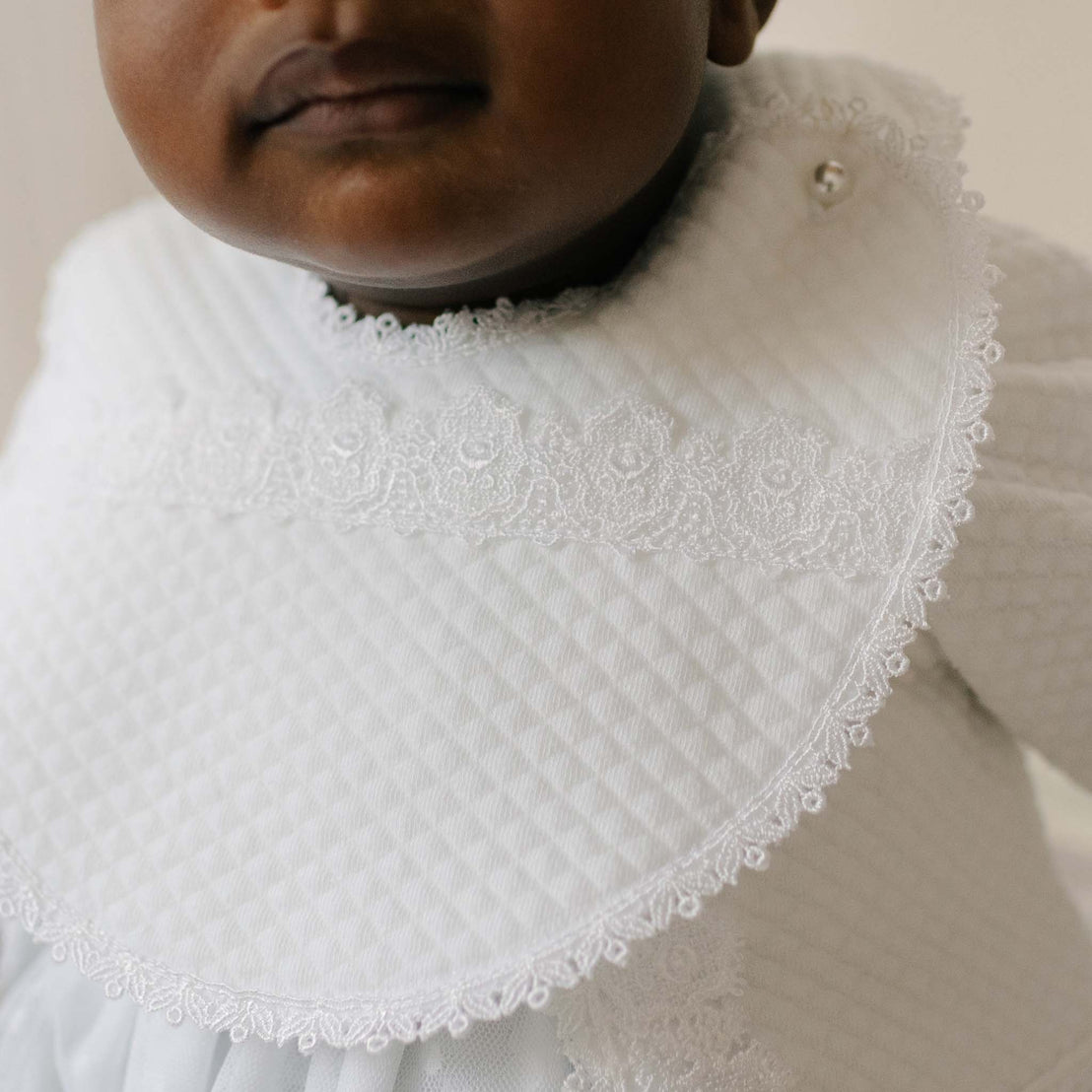 Close-up of the Stella Bib as worn by a baby against a neutral background. Details of the bib feature white Venice lace, edge lace trim, and a pearl style button closure.