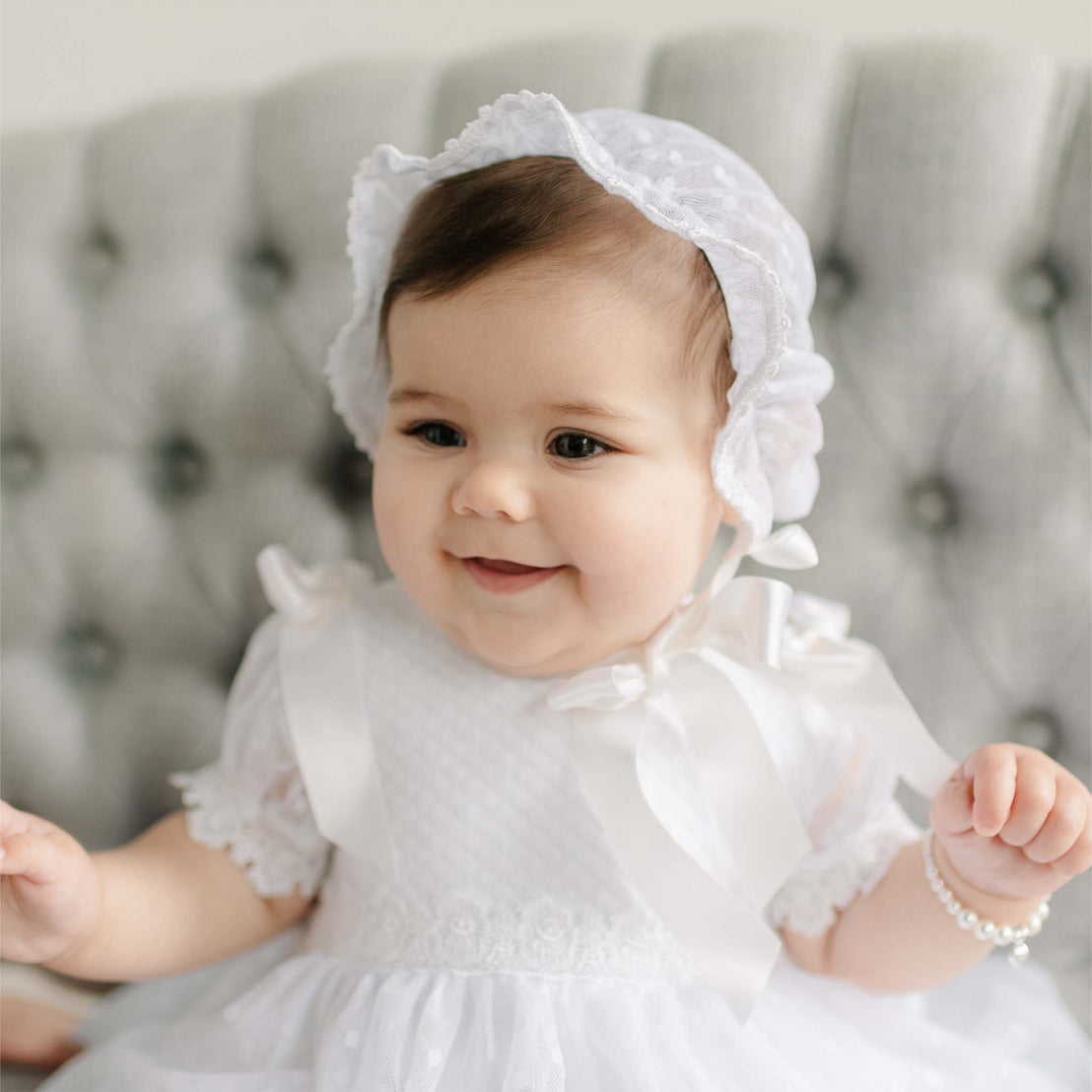 Baby in the Stella Lace Bonnet and matching dress sitting on a gray cushioned chair. Part of Baby Beau & Belle's high end baby clothes.