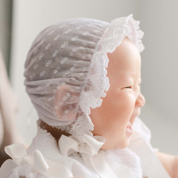 Baby wearing the Stella Lace Bonnet against a soft focus background. The bib features a white dotted lace and a white edge lace trim with silk ribbon ties. Part of Baby Beau & Belle's high end baby clothes.