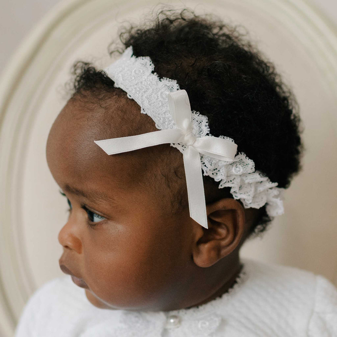 Baby wearing the Stella Lace Headband, looking to the side. Details of the headband include a Venice lace band with elastic and a silk ribbon bow. Part of Baby Beau & Belle's high end baby clothes.